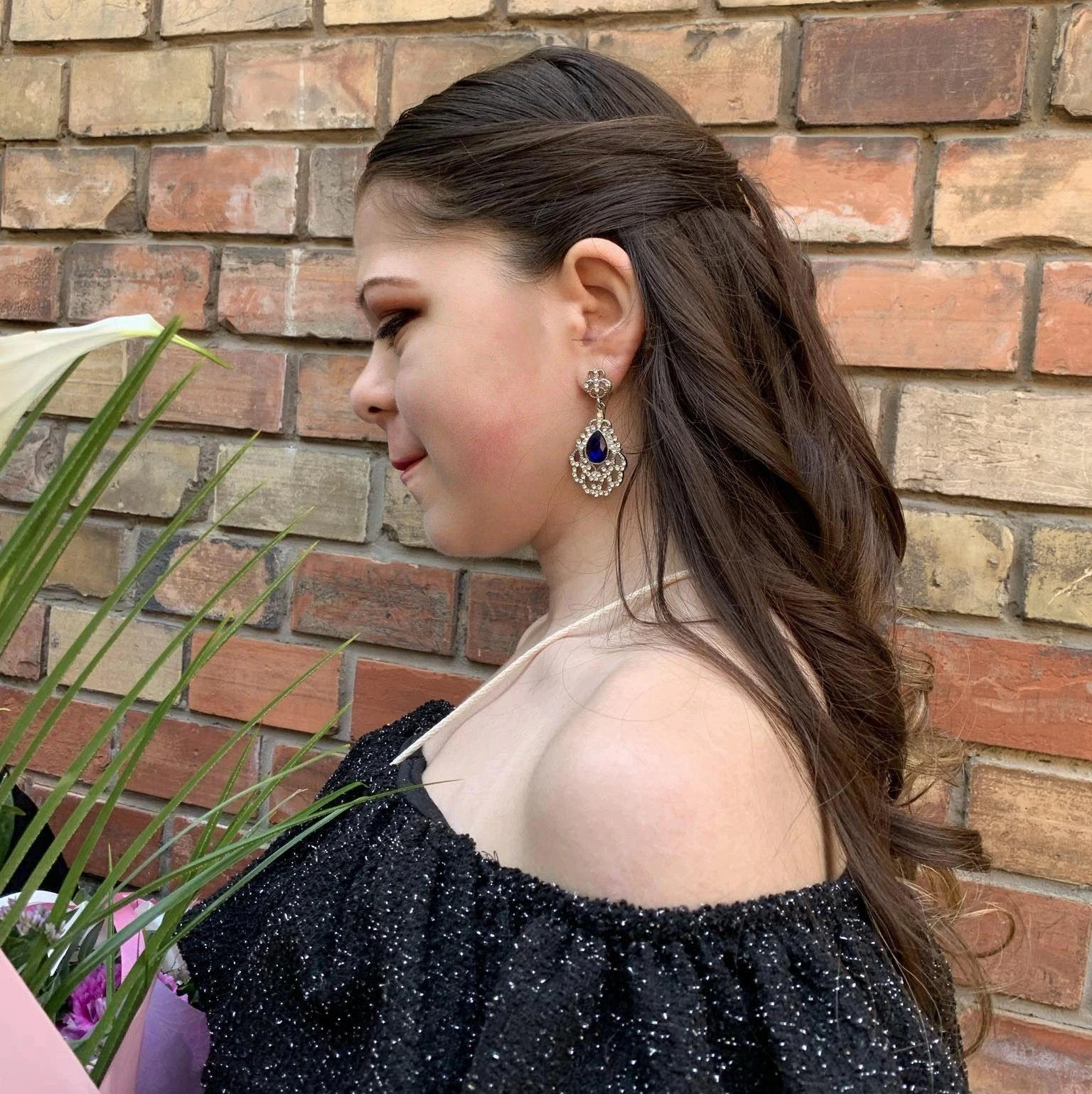 Művészi portré egy sugárzóan elegáns fiatal nőről, akinek
lágy hullámokban omlik le barna haja; az összképet a sötétkék
drágaköves fülbevaló és a vállat szabadon hagyó, éjfekete csillogó
ruha teszi teljessé a rusztikus téglafal előtt.